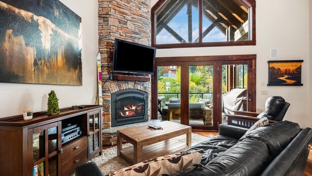 A cozy living room with a stone fireplace, leather sofa, and a television mounted on the wall. - Schweitzer Resort Stay | Deck, Hot Tub & Stunning Views (Sandpoint)