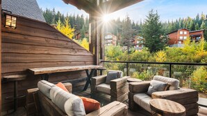 A wooden deck with a bench and seating area, overlooking a forested area with buildings in the background. - Schweitzer Resort Stay | Deck, Hot Tub & Stunning Views (Sandpoint)