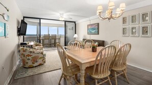 A dining area with a wooden table and chairs, a chandelier, and a view of the ocean through large windows. - Gorgeous 3 Bedroom Condo Oceanfront with Pool! (Ocean City)