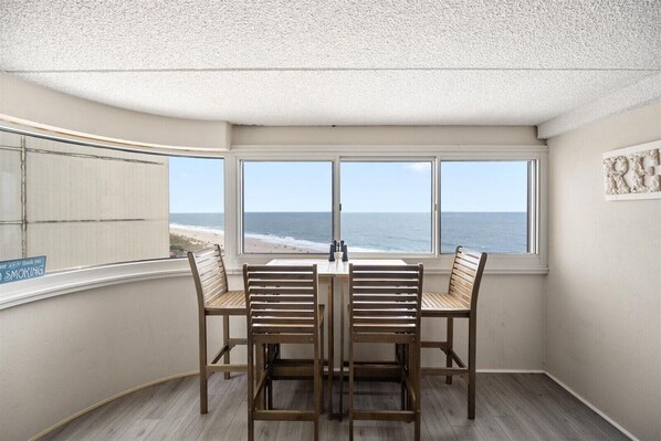 A corner room with a view of the beach, a table with four chairs, and a sign on the window railing.