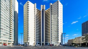 A modern high-rise building with a central atrium, surrounded by a parking lot and other buildings. - Gorgeous 3 Bedroom Condo Oceanfront with Pool! (Ocean City)