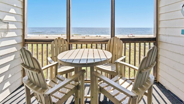 A wooden deck with a table and chairs facing a beach view through large windows.