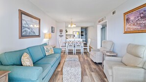 A living room with a teal sofa, beige armchairs, a dining table with chairs, and a kitchen in the background.