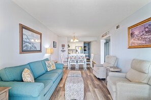 A living room with a teal sofa, beige armchairs, a dining table with chairs, and a kitchen in the background. - Oceanfront Condo with Screened Oceanfront Balcony + Free Attraction Tickets! (North Myrtle Beach)