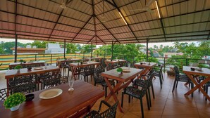 An outdoor restaurant with wooden tables and chairs, a metal roof, and a view of a sports field. - Panakal Residency (Kochi)