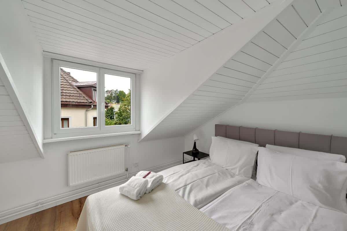 Ein Schlafzimmer mit schrägverdeckter Decke, einem großen Fenster, einem Bett mit weißer Bettwäsche, einem Nachttisch und einem Heizkörper.