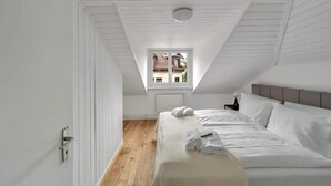Quarto com teto inclinado, janela com vista para uma casa, cama com roupa de cama branca e mesa de cabeceira com luminária.