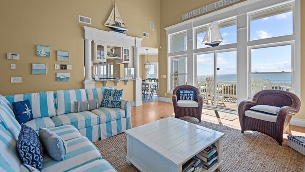 Ein Wohnzimmer mit großem Fenster mit Blick auf den Strand, einem gestreiften Sofa, einem Couchtisch und einem Korbstuhl.