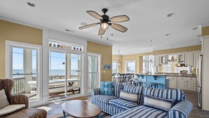 Ein geräumiges Wohnzimmer mit einer großen Couch, einem Couchtisch und einem Deckenventilator. Durch große Glasfenster ist der Meerblick sichtbar.