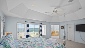 Ein Schlafzimmer mit einem großen Bett, einem Deckenventilator, Blick auf den Ozean durch das Fenster und einem an der Wand befestigten Fernseher.