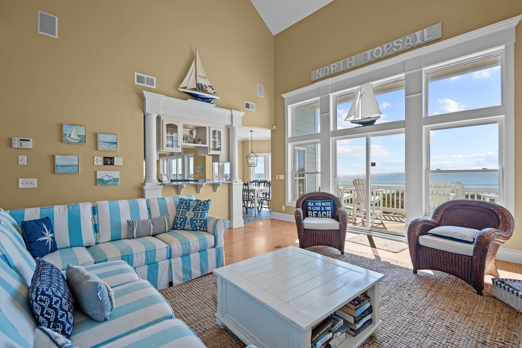 Un salon doté d’une grande fenêtre donnant sur la plage, d’un canapé à rayures, d’une table basse et d’un fauteuil en osier.
