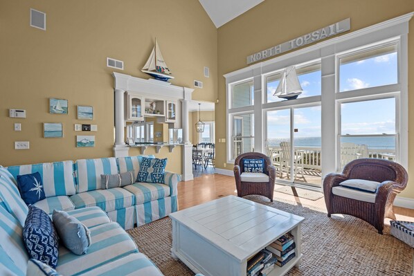 Una sala de estar con un ventanal grande con vista a la playa, un sofá a rayas, una mesa de centro y una silla de mimbre.