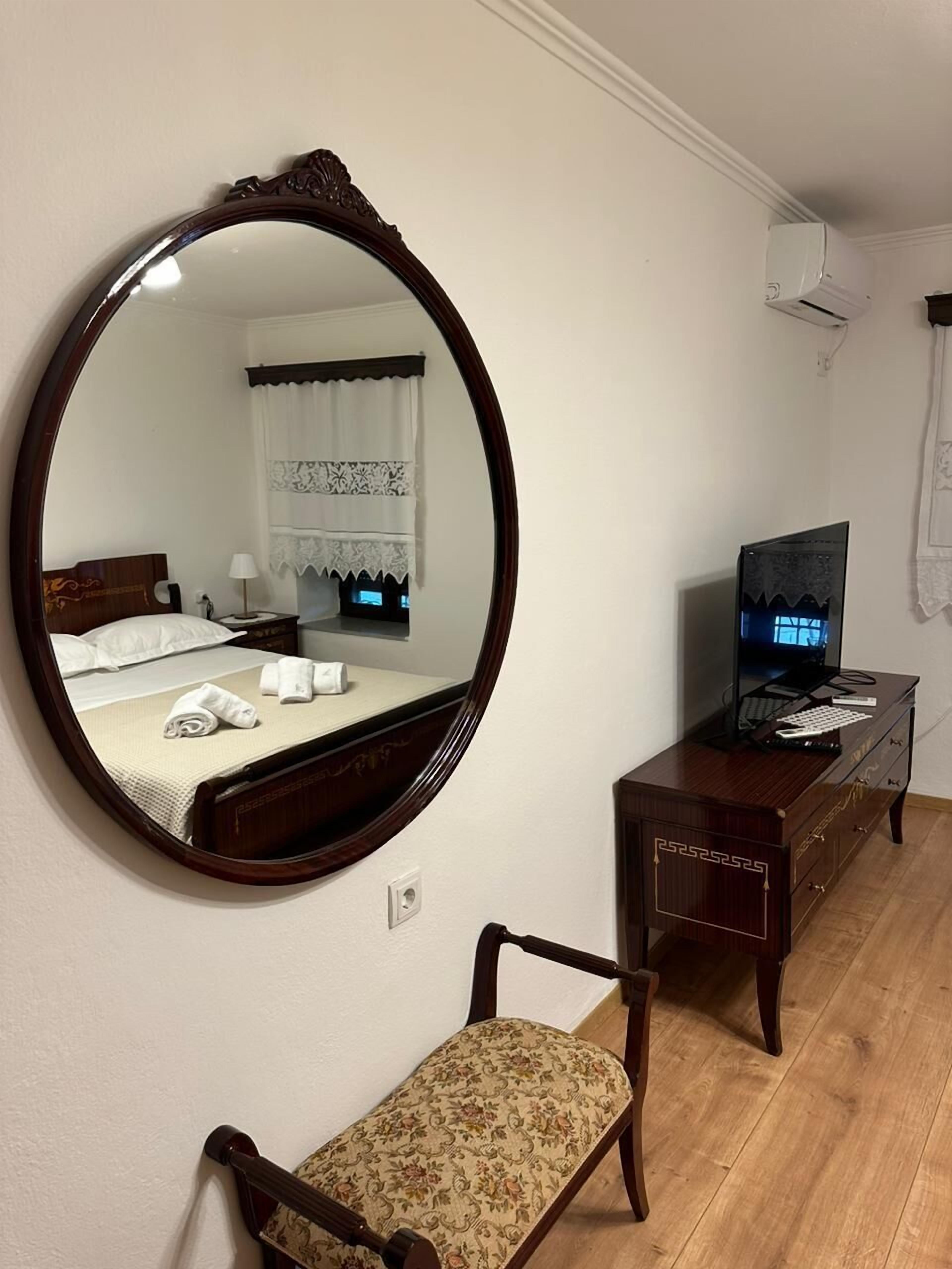 Una habitación de hotel con un espejo redondo, una cama con ropa blanca, un tocador de madera con un televisor y una silla con estampado floral.