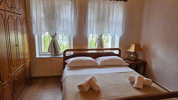A bedroom with a wooden bed, two windows with white curtains, a bedside lamp, and a wooden wardrobe.