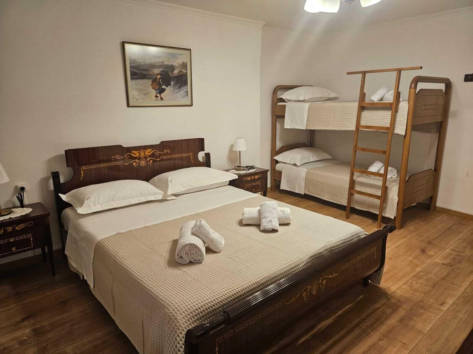 A bedroom with a large bed, a smaller bunk bed, a nightstand, a bedside table, and a wall-mounted painting.