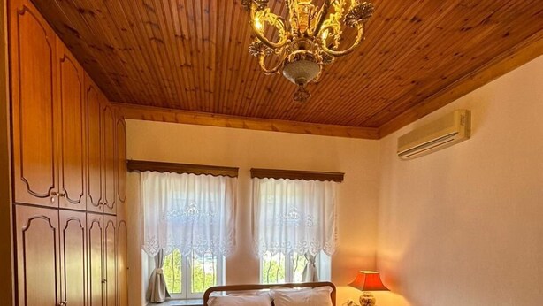 Une chambre à coucher avec un plafond en bois, un lustre, un lit recouvert de draps blancs, deux fenêtres avec des rideaux et une table de chevet avec une lampe.