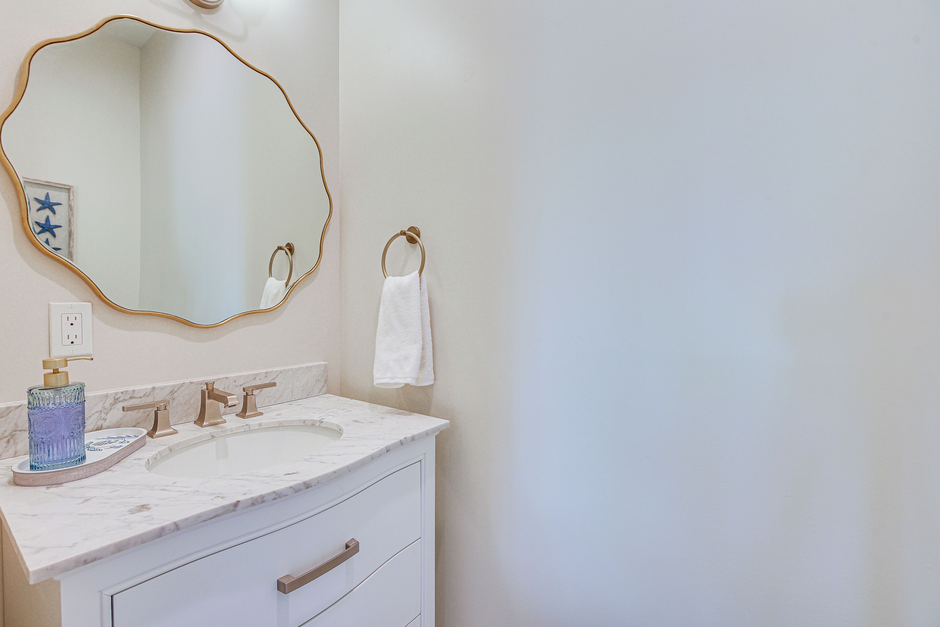 A bathroom with a large oval mirror, a white sink with a marble countertop, and a gold faucet.