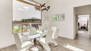 Ein moderner Essbereich mit einem Glastisch und weißen Stühlen, einer dekorativen Leuchte sowie Blick ins Freie.