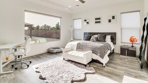 Ein Schlafzimmer mit einem großen Bett, einem Schreibtisch und einem Stuhl. Es gibt einen Deckenventilator, ein Fenster und einen Nachttisch mit einer Lampe.