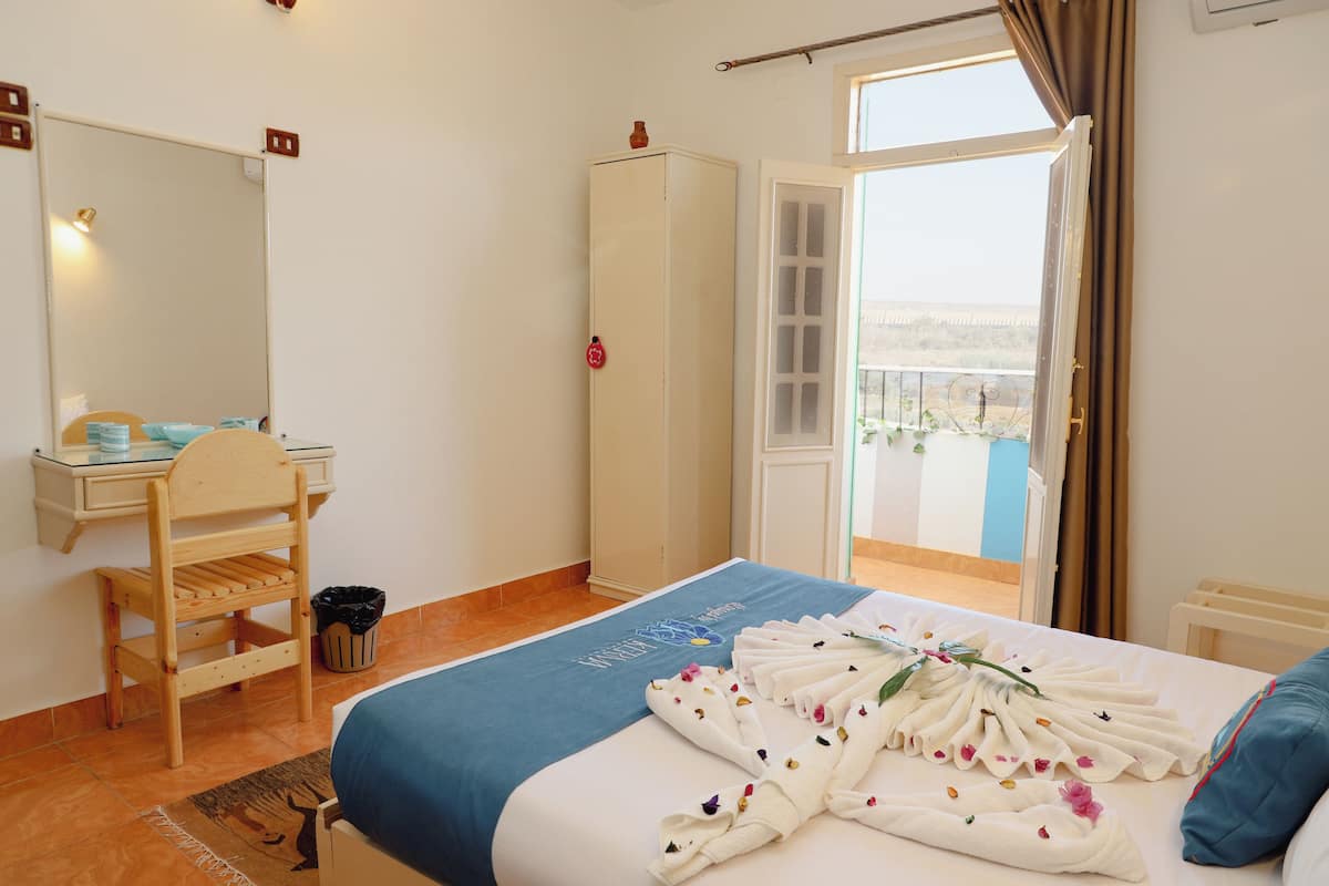 A hotel room with a bed, a wooden chair, a desk, a wardrobe, and a balcony with a view.