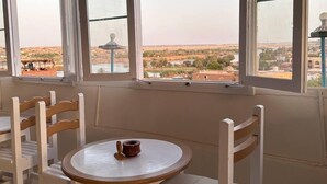 A room with a round table and white chairs, large windows with a view of a landscape, and a small decorative item on the table. - Ekadolli Nubian house - by kerma hospitality (Aswan)