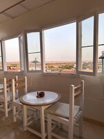 Una habitación con una mesa redonda y sillas blancas, ventanas amplias con vistas a un paisaje y un pequeño adorno sobre la mesa.