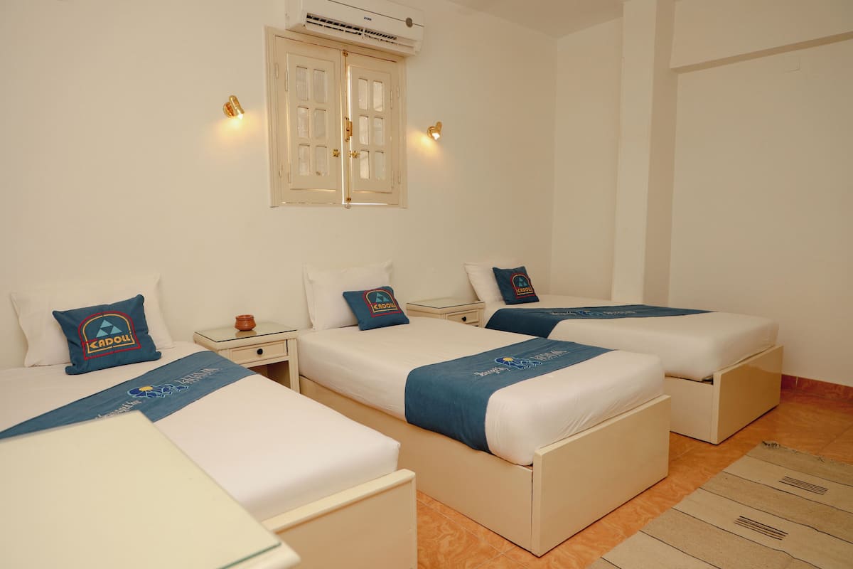 A hotel room with three single beds, each with a blue bedspread and a pillow with a logo. There is a nightstand between the beds, a small table with a lamp, and a window with a curtain.