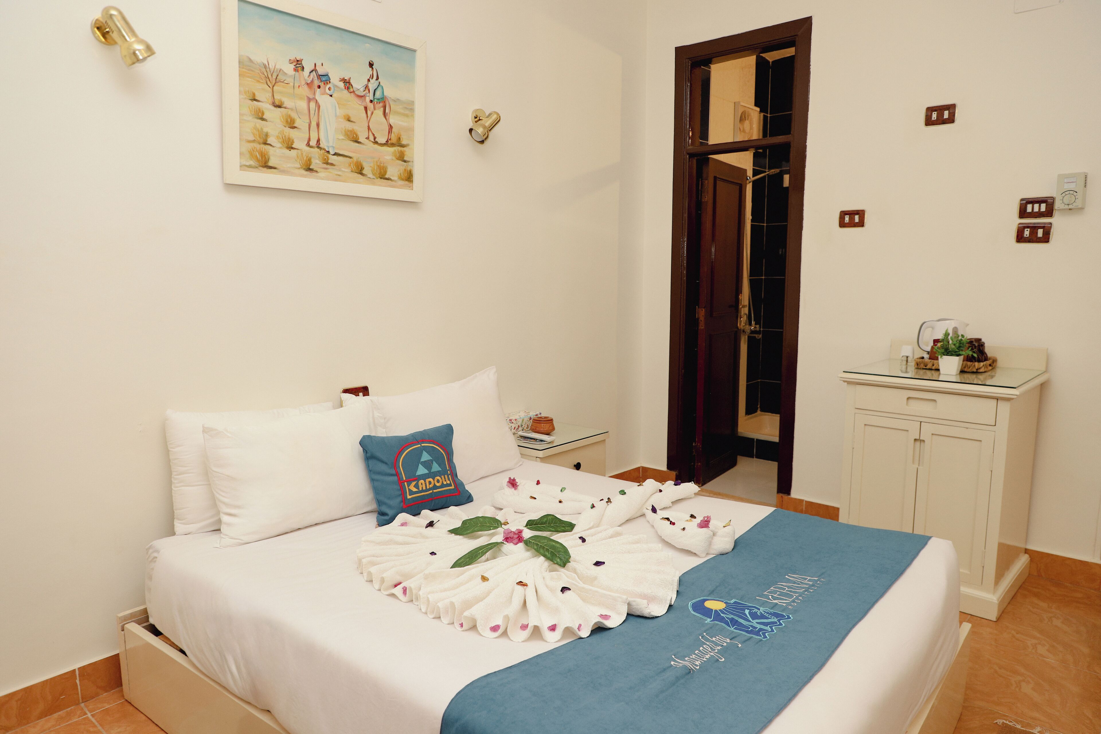 A neatly made bed with a towel arrangement, a painting on the wall, and a small bedside table with a lamp.