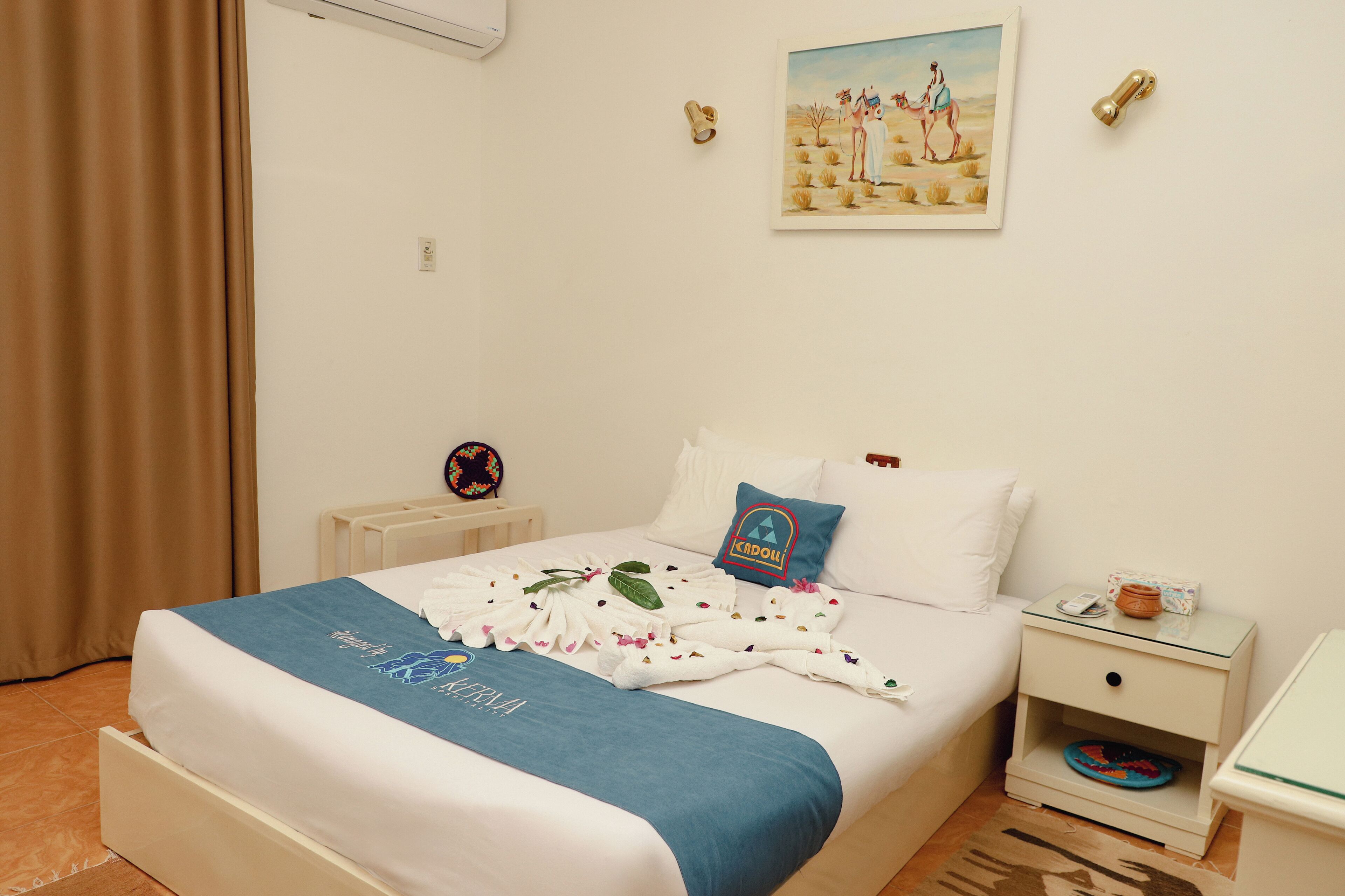 Un lit bien fait, avec une serviette bleue, des oreillers blancs et un coussin décoratif orné d’un logo. Une petite table de chevet, sur laquelle se trouvent un téléphone et un objet décoratif, est placée à côté du lit.