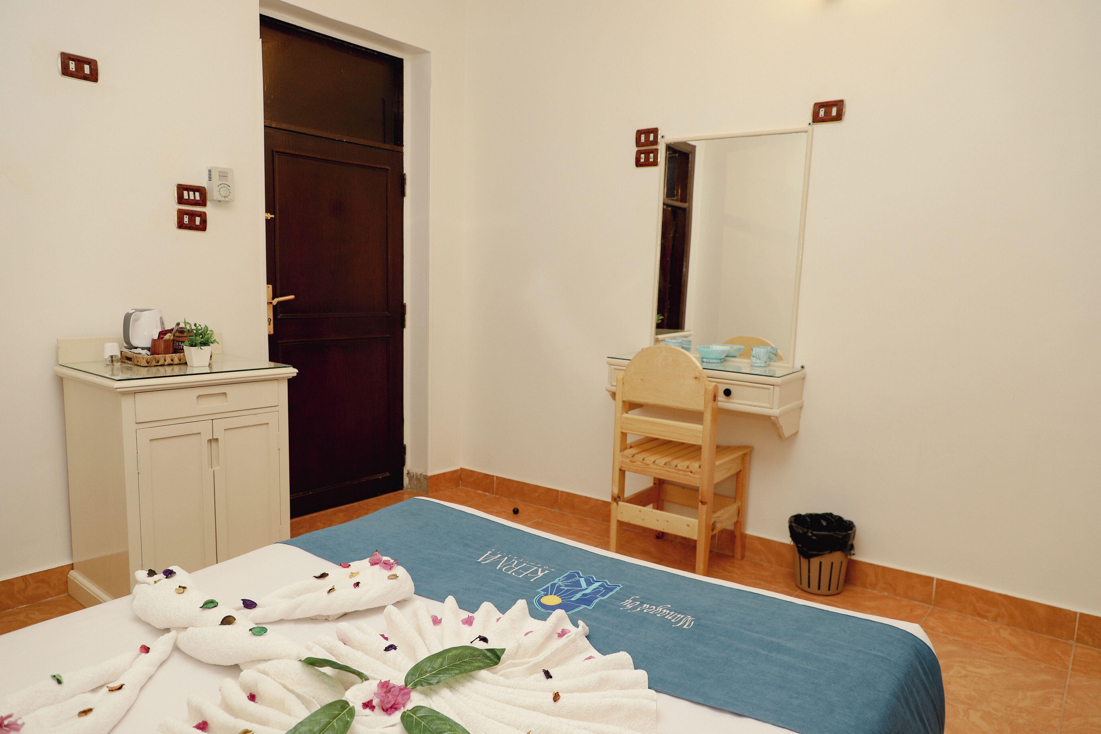A hotel room with a bed, a wooden chair, a small table with toiletries, a mirror, and a trash bin.