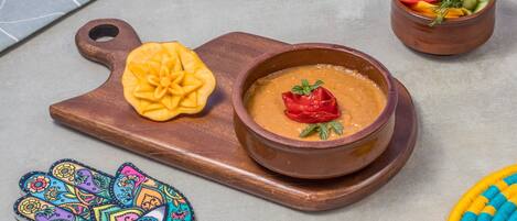 A wooden serving board with a bowl of soup garnished with a red chili pepper, a carved pumpkin, and a small bowl of assorted vegetables.