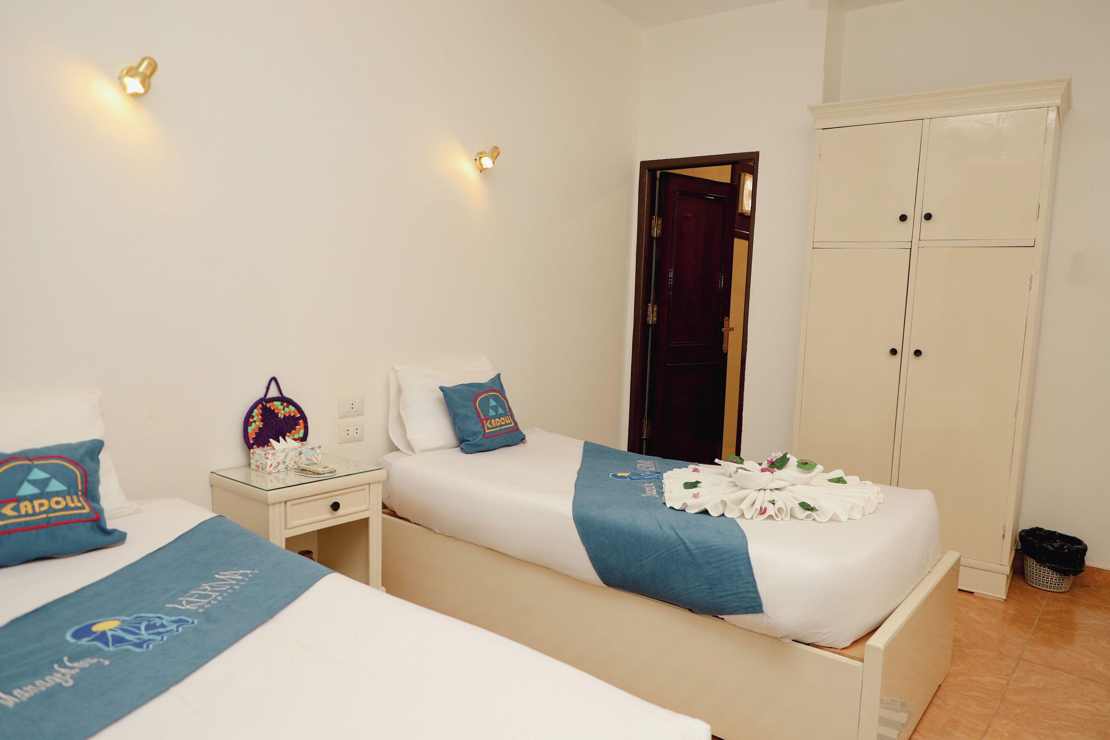 A hotel room with two beds, a wardrobe, a nightstand, and a wall-mounted light fixture.