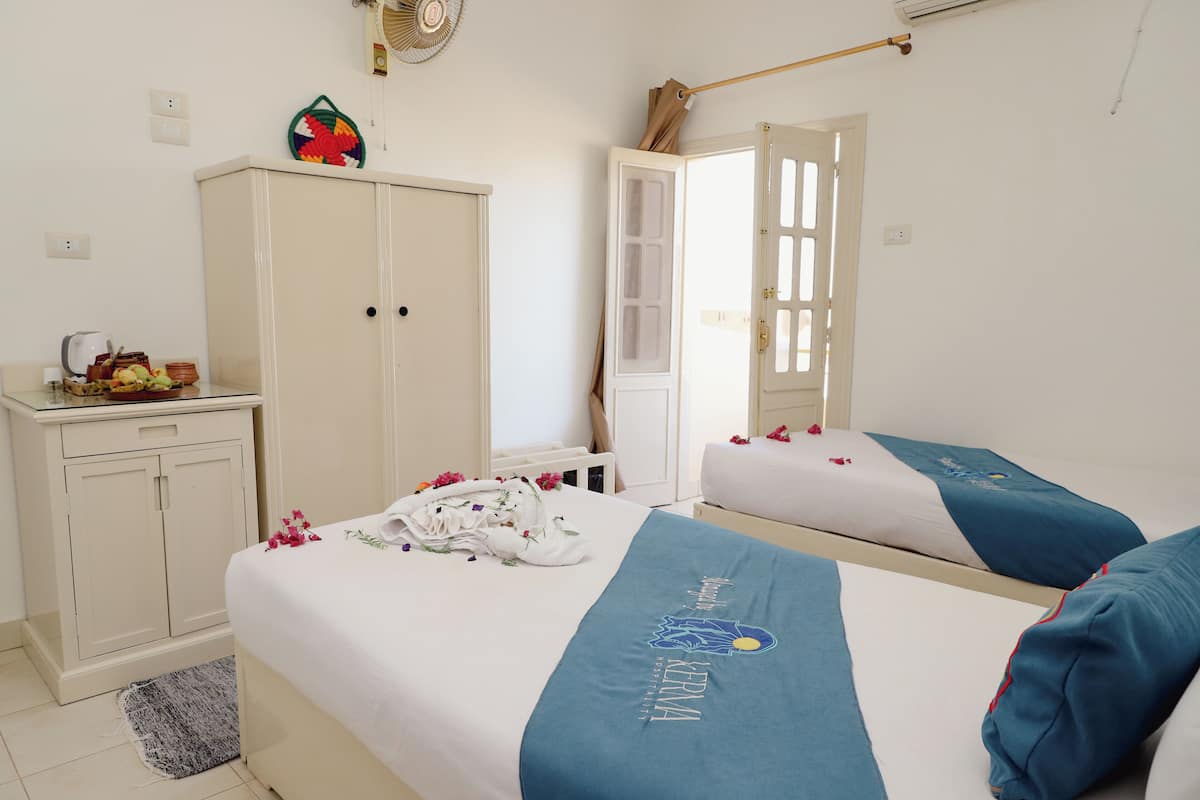 A hotel room with two beds, a wardrobe, a desk with a kettle, and a decorative wall hanging.