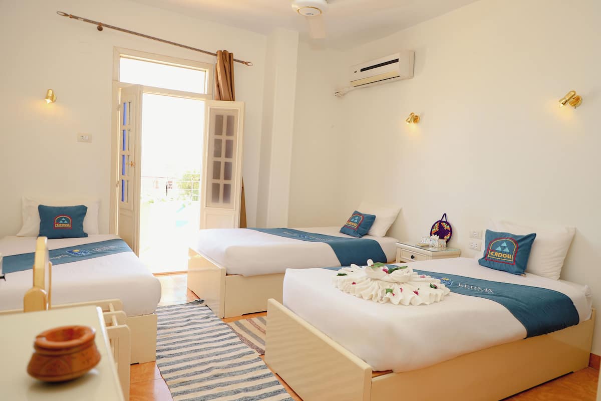 A hotel room with two beds, a small table, a chair, a window with curtains, an air conditioning unit, and wall-mounted lights.