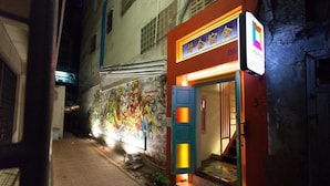 A narrow alley with a brightly lit building featuring a colorful mural on the wall. - Stash Hostel (Bangkok)