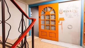 A hotel entrance with a red and blue door, a staircase with a red handrail, and a wall with a 'KUVAIN' logo. - Stash Hostel (Bangkok)