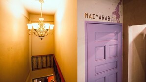 A hallway with a chandelier, a purple door labeled "MAIYARAB," and a staircase with a red railing. - Stash Hostel (Bangkok)