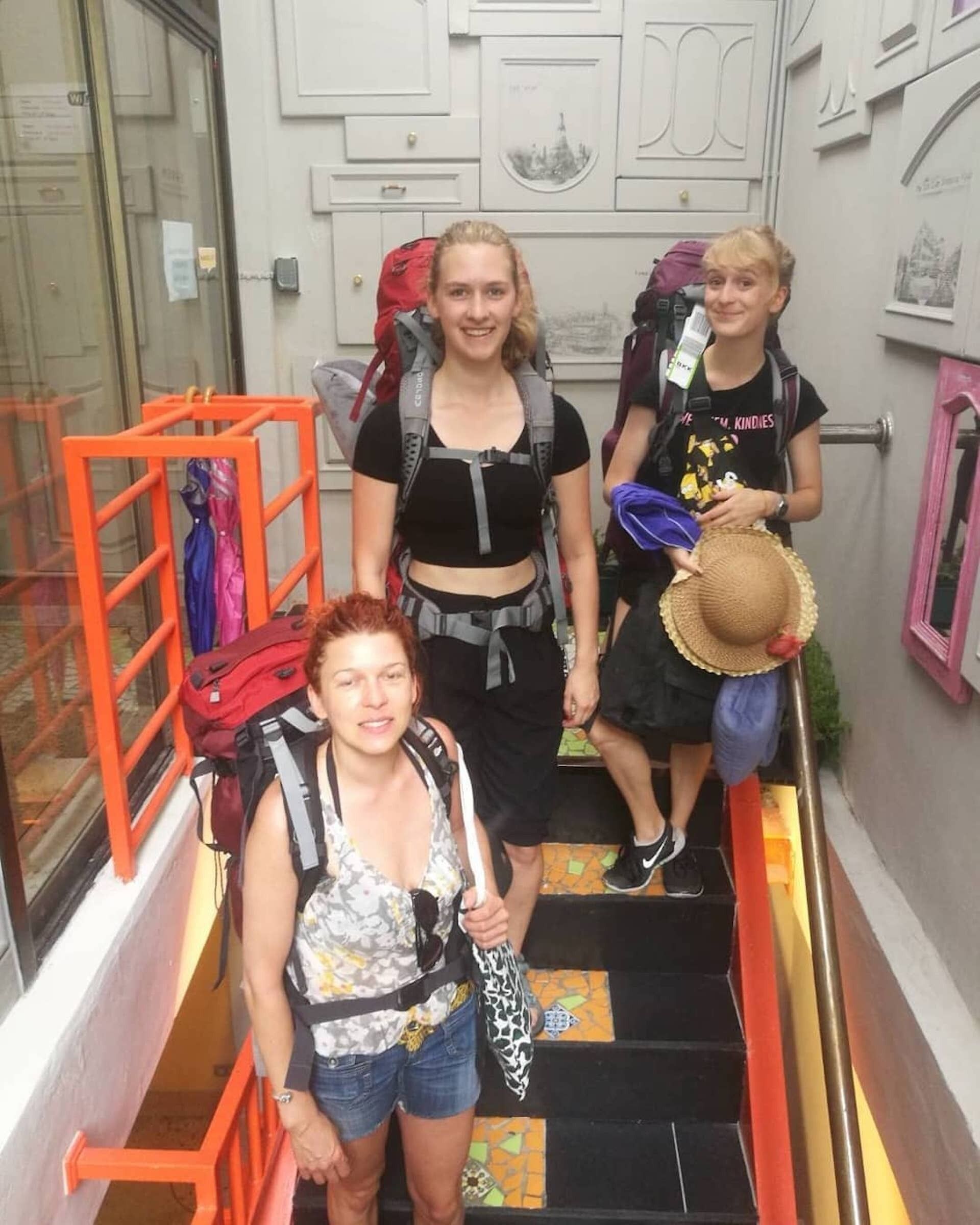 Three people ascending a narrow staircase with orange railings, carrying backpacks.