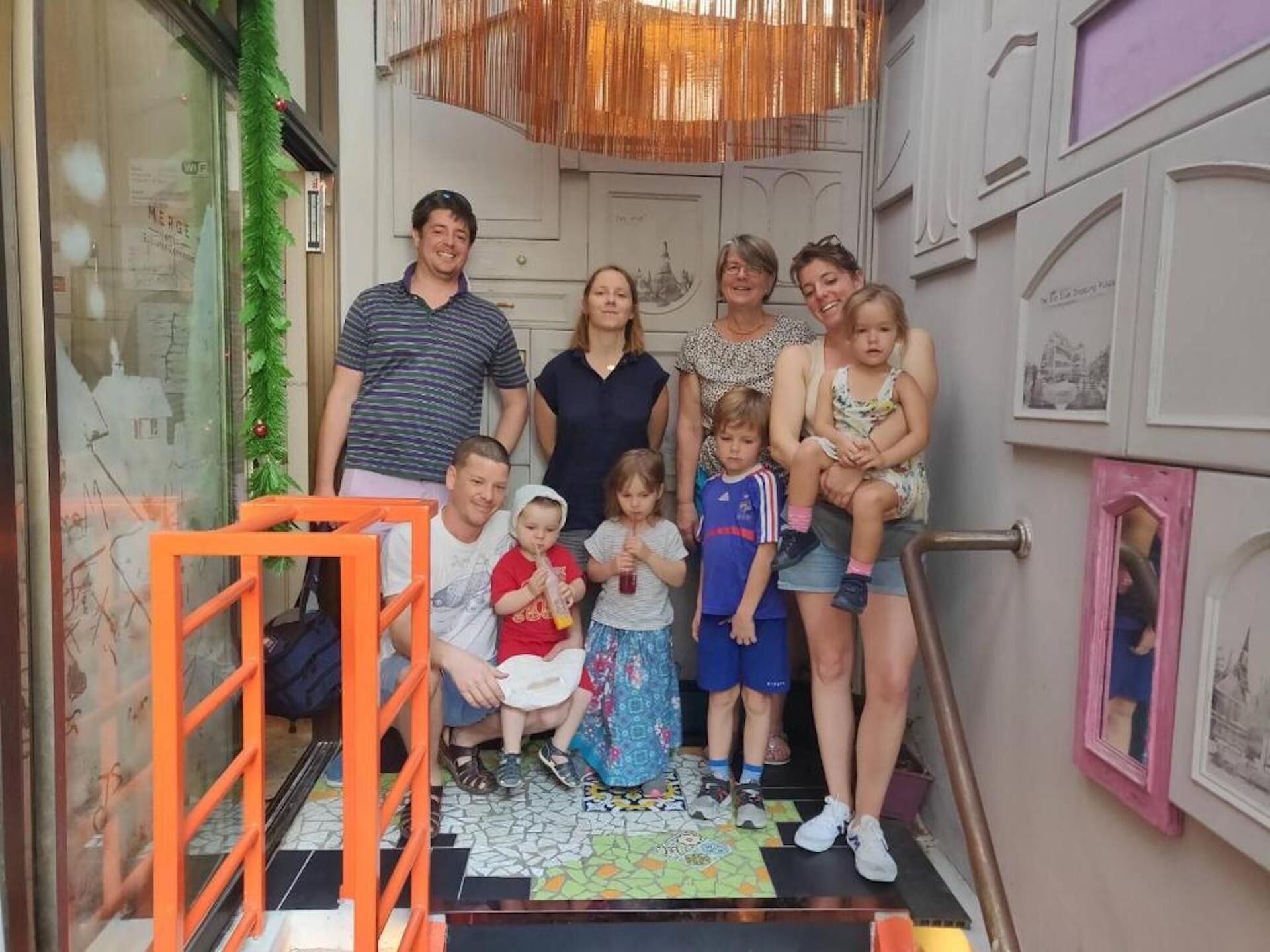 A family group photo in a hallway with a staircase, a decorative wall, and a door with a window.
