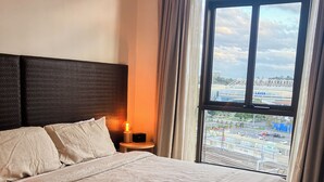 A hotel room with a large bed, a headboard, a bedside table with a lamp, and a view of the city through a window. - Jolimont Views - Centrally located top floor apt overlooking MCG/Rod Laver (East Melbourne)