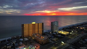 A high-rise hotel with multiple floors and a parking garage, overlooking a beach and ocean at sunset. - Treasure Island 804- Heated Pool, Hot Tub, Grills, FREE Golf & Beach Service! (Panama City Beach)