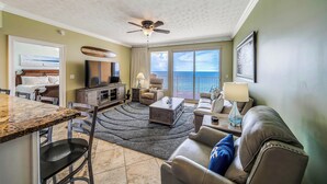 A living room with a large window, a ceiling fan, and a dining area with a granite countertop. - Treasure Island 804- Heated Pool, Hot Tub, Grills, FREE Golf & Beach Service! (Panama City Beach)