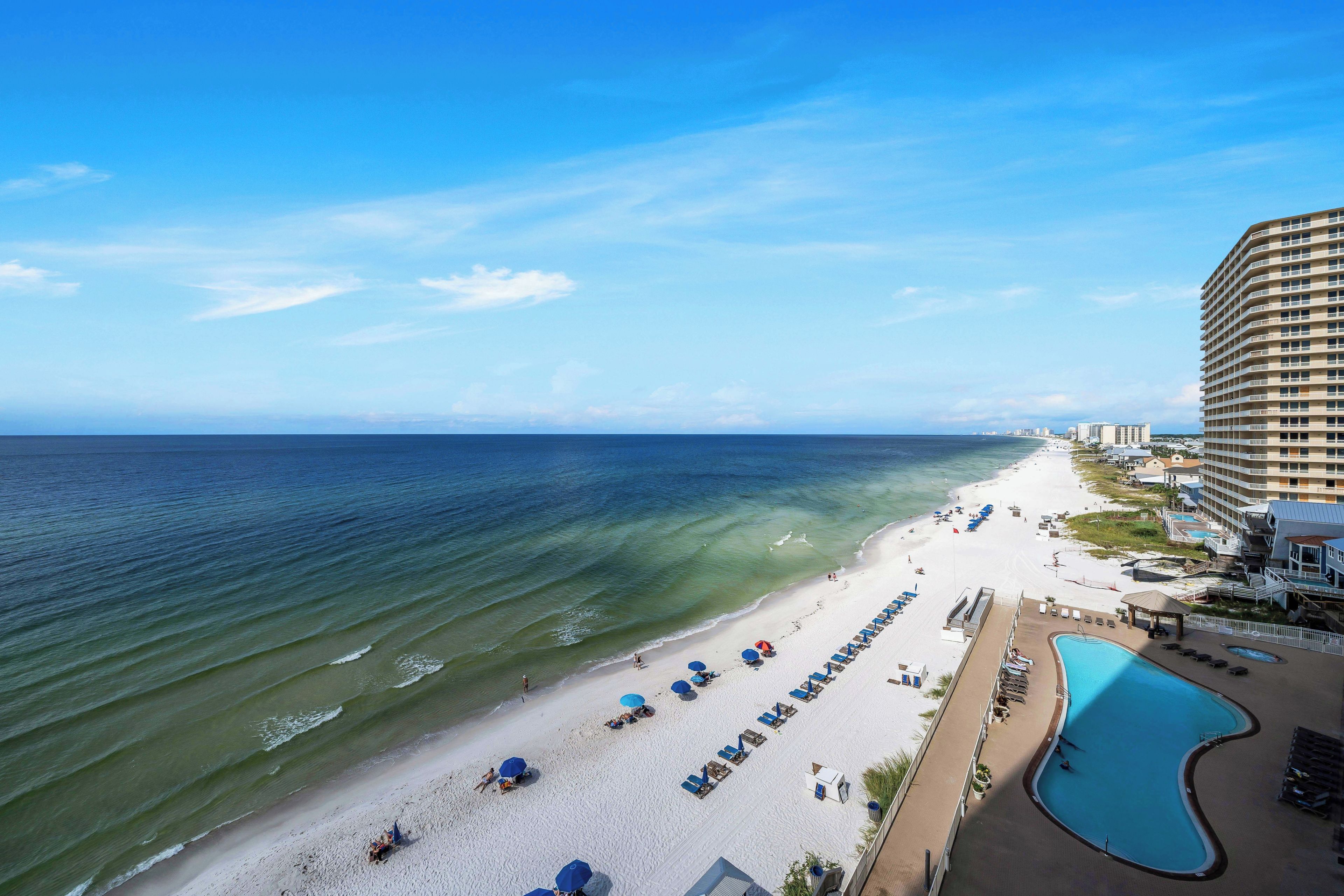 A beachfront hotel with a pool, beach chairs, umbrellas, and a view of the ocean.