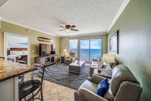 A living room with a large window, a ceiling fan, and a dining area with a granite countertop. - Treasure Island 804- Heated Pool, Hot Tub, Grills, FREE Golf & Beach Service! (Panama City Beach)