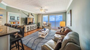 A spacious living room with a dining area, a ceiling fan, a large window with a view, and a balcony. - Pet Friendly Treasure Island 1504- Heated Pool, Gym, FREE Golf &Beach Service (Panama City Beach)