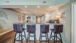 A kitchen with a central island, bar stools, and a view into the dining area. - Pet Friendly Treasure Island 1504- Heated Pool, Gym, FREE Golf &Beach Service (Panama City Beach)