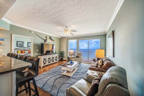 A spacious living room with a dining area, a ceiling fan, a large window with a view, and a balcony. - Pet Friendly Treasure Island 1504- Heated Pool, Gym, FREE Golf &Beach Service (Panama City Beach)