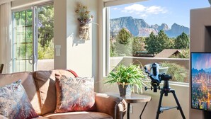 Ein Wohnzimmer mit großem Fenster und Blick auf die Berge, einem auf einem Stativ stehenden Teleskop und einem Blumentopf auf einem kleinen Tisch.