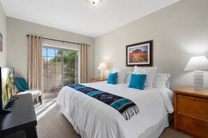 A hotel room with a bed, two bedside lamps, a desk with a chair, a large window with curtains, and a framed picture on the wall.