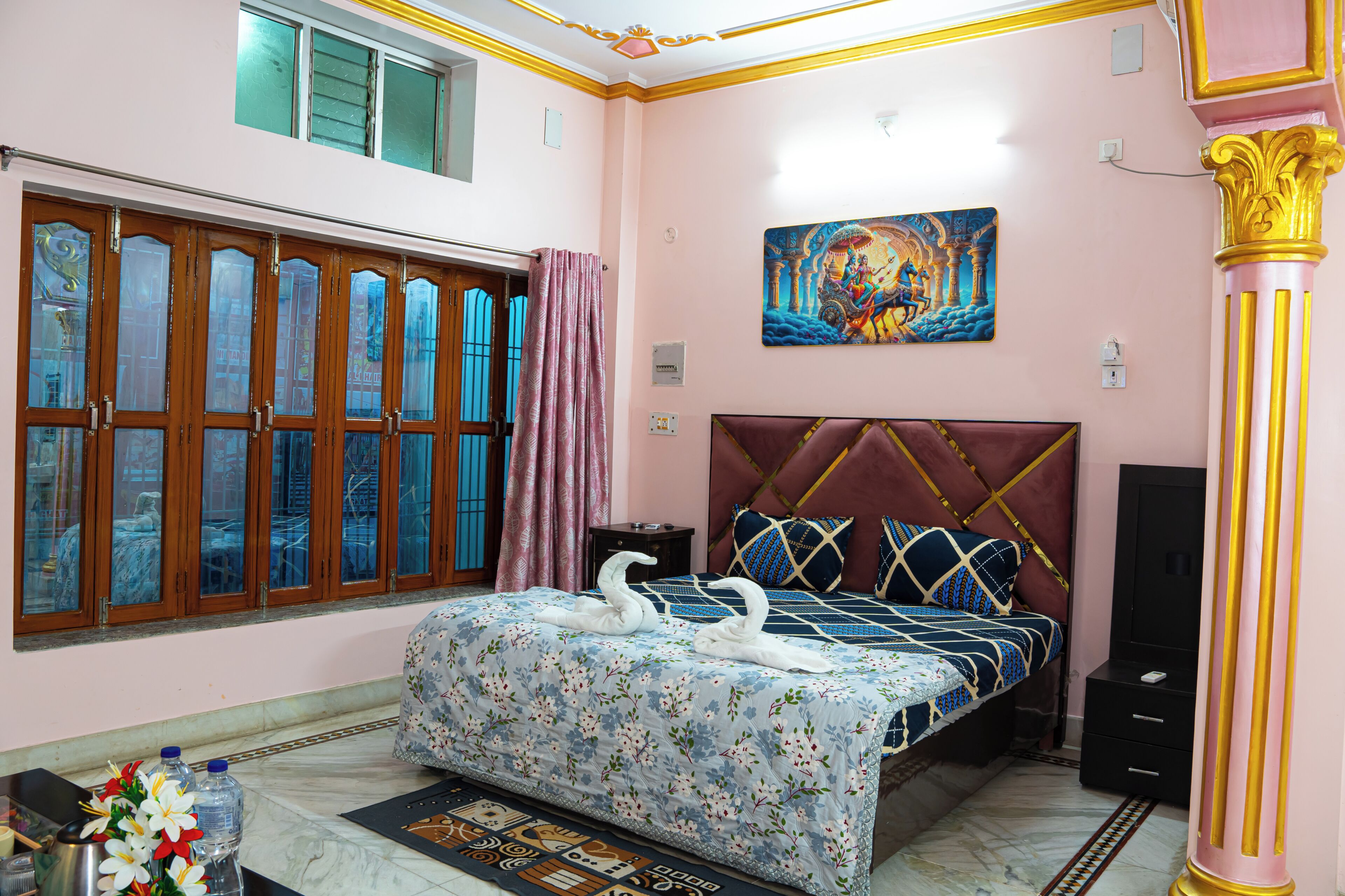 A bedroom with a bed, a window with curtains, a painting on the wall, and a decorative column.
